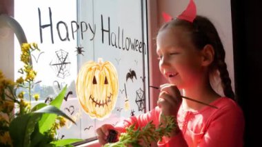 Pencerede balkabağı boyayan şeytan boynuzlu çocuk Cadılar Bayramı 'nı kutluyor. Küçük çocuk oda dekore ediyor. Kağıt yarasalar sonbahar tatilini kutluyor. Yaratıcı eğlence, yeni gerçeklik.