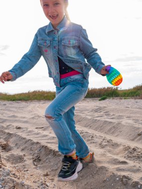 Mutlu çocuk, boş sonbahar kumsalında zıplayarak eğleniyor. Sarışın kız beyaz kum yolunda yürüyor. Mavi gökyüzü arka planı. Yaşam tarzı fotoğrafı. Parlak gökkuşağı renginde neşeli çocuk patlat onu oyuncak.