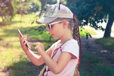 Beyzbol şapkalı, uzun örülü saçları ve açık hava güneşli bir günü olan sevimli küçük bir kız. Gençlik kültürü yaşam tarzı. Yeşil çimlerde mesajlaşan mutlu gezgin çocuk. Otantik okul çocuğu yürüyor.