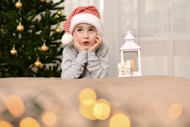 Karnaval şapkalı bir çocuk şaşkınlıkla gözlerini yuvarladı ve elleriyle yüzünü sıktı. Noel ağacının yanında otururken.