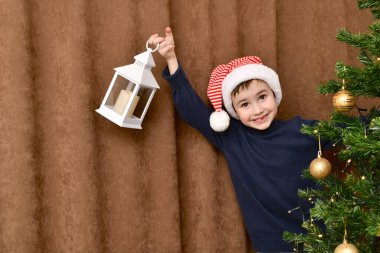 Smiling kid, with an enthusiastic face, in a striped Santa Claus cap, suddenly looked out from behind the Christmas tree with a lantern, in a high pitched hand. clipart