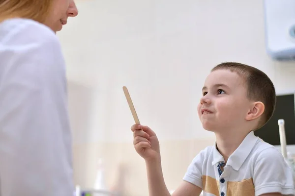 医者と患者は子供です その少年はへらを手に持ち 賢く笑っているように見える 診療所の事務所で — ストック写真