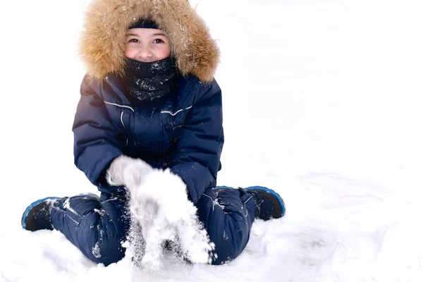 Uma criança em um macacão com uma balaclava de um fole. Sentado em um dia de inverno, muita neve com as mãos em mitenes. Imagens Royalty-Free