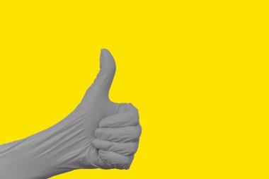 Elinin kapalı bir yumruğuyla, başparmağı yukarı doğru uzatılmış, koruyucu gerilmiş bir nitril eldiven giymiş, nihai gri, arka planda ışık saçan sarı üniformalı bir jest..