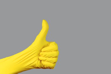 Elinin kapalı bir yumruğuyla baş parmağı yukarı doğru uzamış, koruyucu gerilmiş bir nitril eldiven giymiş, sarı ışıklı, tek bir gri arka plana karşı..