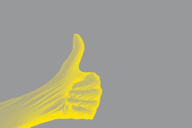 Bir elin sıkılmış bir el hareketi baş parmağı yukarıya doğru uzanmış koruyucu gerilmiş nitrile eldiven üzerinde sarı ışıklandırılmış tek bir gri arka planda. Gri renkli gölgelerle.
