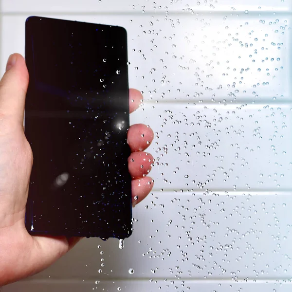 Telefon komórkowy w ręku pod kroplami wody na tle białej płytki. Pranie wodoodpornego smartfona. — Zdjęcie stockowe