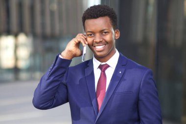 Resmi takım elbiseli, cep telefonuyla konuşan mutlu genç iş adamının portresi. Siyah Afrikalı Afro-Amerikalı adam arıyor, akıllı telefonuyla konuşuyor ve gülümsüyor.