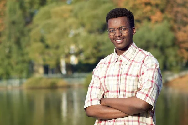 Porträt eines gut aussehenden schwarzen Afroamerikaners, der im Hemd in einem Park am See steht und mit verschränkten Händen lächelt. Ethischer Student. — Stockfoto