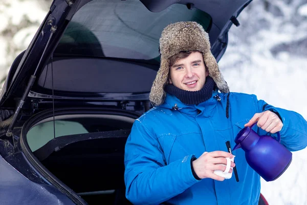 Handsome young man in warm winter clothes is drinking hot drink tea or coffee from a thermos sitting in the trunk of a car and smiling. Vacation, traveling by car, snowy cold weather.