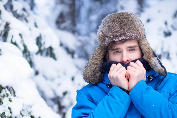 寒い天候に苦しんで耳フラップ付きの帽子の冷凍男。暖かい服、震え、雪の背景に木の冬の雪の森の中の若い男. — ストック写真