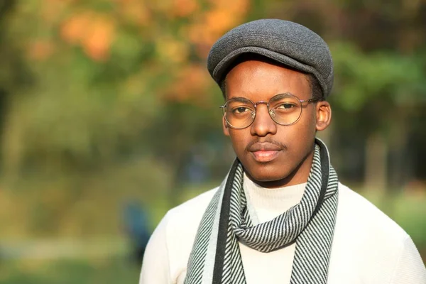 Porträtt av allvarlig pensiv stilig svart afrikansk afro amerikansk ung man i elegant fashionabla hatt och halsduk i glasögon tittar på kameran, promenader i gyllene höstpark. — Stockfoto