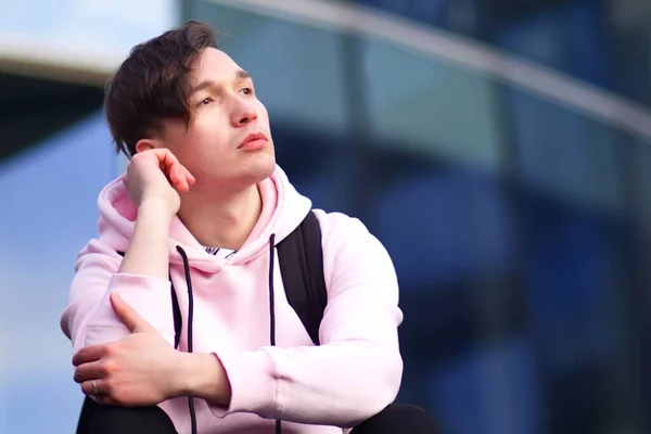 Pensamiento pensativo joven triste está sentado al aire libre y mirando a la distancia. Molesto melancólico brooding chico está pensando en el futuro en sudadera con capucha rosa con mochila — Foto de Stock