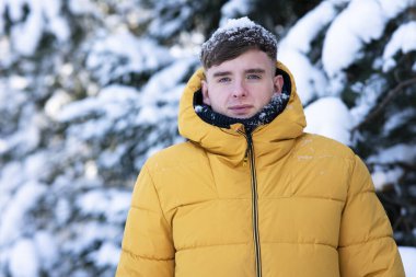 Yakışıklı genç bir adamın portresi açık havada sıcak sarı ceketle yürüyor, adam karlı bir günde kış ormanında kameraya bakıyor.