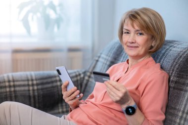 Evde cep telefonu, akıllı telefonu olan, satın alan, kullanan, internet alışverişi için kredi kartını elinde tutan, gülümseyen mutlu, mutlu yaşlı bir kadın var. Çevrimiçi ödeme kavramı. 