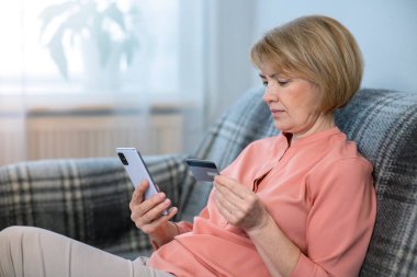 Evde cep telefonu, akıllı telefonu olan, satın alan, kullanan, internet alışverişi için kredi kartını elinde tutan, ciddi şekilde odaklanmış yaşlı, güzel bir kadın. Çevrimiçi ödeme kavramı. 