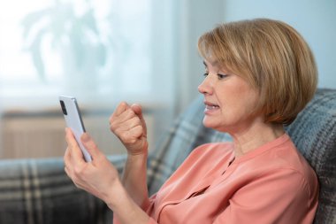 Öfkeli, öfkeli, sinirli bir kadın, yaşlı, yetişkin bir bayan cep telefonuyla ekranda yumruğunu sallıyor. Arızalı akıllı telefondan kaynaklanan sorunlar. Olumsuz konuşma. 