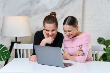 İki güzel, mutlu, pozitif kadın arkadaşın portresi, iş arkadaşları projede birlikte çalışıyorlar, ekrana bakıyorlar, dizüstü bilgisayar kullanıyorlar, gülümsüyorlar, gülüyorlar, evde komik içerikler izliyorlar.