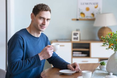 Yakışıklı, mutlu, pozitif bir adamın portresi. Mutfaktaki masada oturan, sabahın köründe pijamada kahve ya da çay içen, kahvaltı yapan, kafeini seven, kameraya bakan genç adam.