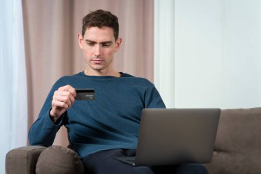 Yakışıklı, ciddi bir adamın portresi, dizüstü bilgisayarı olan, internet mağazasından sipariş veren, evdeki kanepe veya kanepede oturan banka kartına bakan genç çekici bir adam. Çevrimiçi alışveriş kavramı