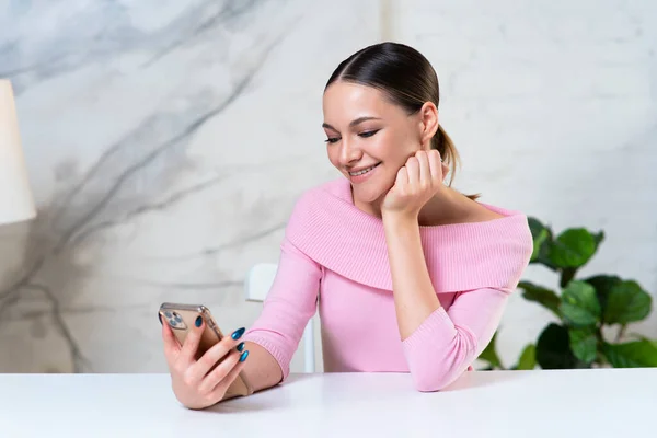 Retrato de menina positiva feliz, jovem mulher bonita olhando para a tela de seu telefone celular, usando gadget smartphone em casa na mesa e sorrindo. — Fotografia de Stock