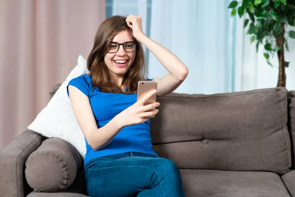 Πορτρέτο του χαρούμενου θετική ενθουσιασμένοι κορίτσι, νεαρή επιτυχημένη όμορφη γυναίκα με γυαλιά χαμογελά, γελώντας, κοιτάζοντας την οθόνη του κινητού τηλεφώνου της, χρησιμοποιώντας το smartphone, γιορτάζει την επιτυχία κερδίσει στο σπίτι — Φωτογραφία Αρχείου