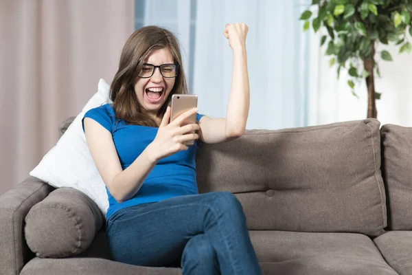 Πορτρέτο του χαρούμενου χαρούμενη ενθουσιασμένος κορίτσι, νεαρή επιτυχημένη γυναίκα κοιτάζοντας την οθόνη του κινητού τηλεφώνου της, γιορτάζει την επιτυχία, νίκη, καλά νέα στο smartphone στο σπίτι στον καναπέ, καναπέ, ουρλιάζοντας — Φωτογραφία Αρχείου