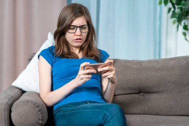 Güzel, neşeli bir genç kız akıllı telefon kullanıyor, gözlüklü genç bir kadın cep telefonuyla ilginç bir oyun oynuyor, evinde faiziyle, oturma odasındaki kanepede oturuyor.