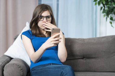 Şok olmuş genç kızın portresi, korkmuş stresli genç kadın cep telefonuna bakıyor, olumsuz haberler okuyor, akıllı telefondan mesaj okuyor, kanepede oturuyor, evdeki kanepede oturuyor.