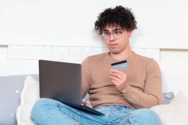Yakışıklı, rahat bir adamın portresi, gözlüklü, dizüstü bilgisayarlı, evde yatak odasında kredi kartıyla internet mağazasından sipariş veren Latin bir genç. Çevrimiçi alışveriş kavramı