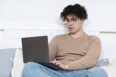 Serbest çalışan ciddi bir etnik erkek portresi, gözlüklü yakışıklı Latin bir adam yatak odasında yatıyor ve bilgisayarı evde kullanıyor. Uzak çalışma, iş, öğrenme, eğitim.