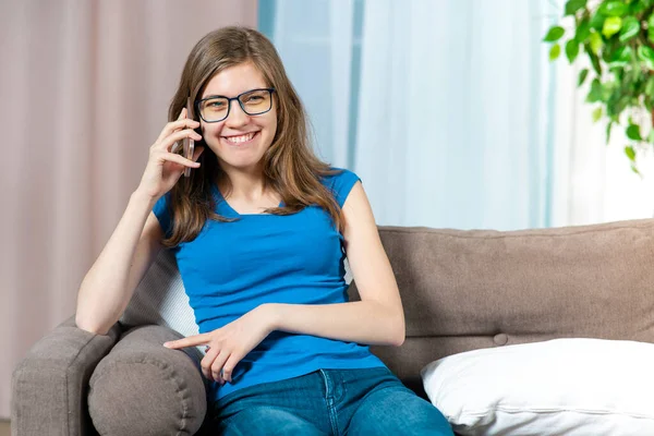 Πορτρέτο του ευτυχισμένου θετική χαρούμενο κορίτσι, νεαρή όμορφη γυναίκα με γυαλιά χαμογελά, γελώντας, μιλώντας στο κινητό τηλέφωνο, καλώντας, έχοντας συνομιλία στο smartphone, κουβεντιάζοντας στον καναπέ στο σπίτι — Φωτογραφία Αρχείου