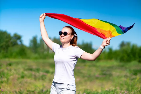 Retrato de mujer joven lesbiana en gafas de sol, hermosa chica está ondeando arco iris LGBT color gay bandera en verano día soleado sobre fondo natural. Comunidad LGBT, concepto de igualdad de relaciones homosexuales — Foto de Stock