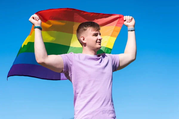 Retrato de joven feliz orgulloso hombre gay, chico guapo está ondeando arco iris LGBT color gay bandera en verano día soleado sobre fondo natural. Comunidad LGBT, concepto de igualdad de relaciones homosexuales — Foto de Stock