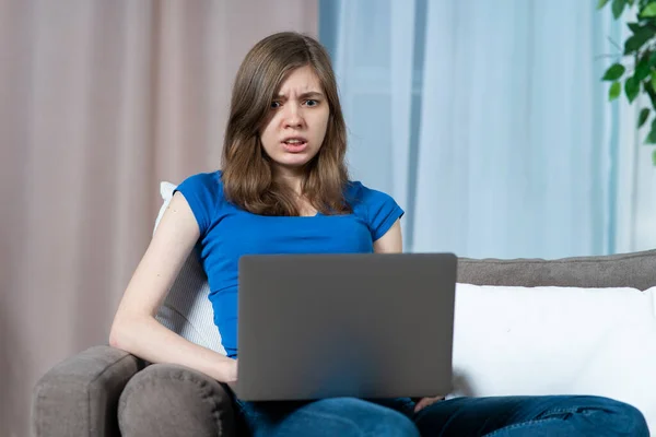 Portrait of scared stressed frighten teen girl, young afraid beautiful woman looking at screen of her laptop computer with fear, sitting it home on sofa or couch. Internet fraud concept. Stock Photo