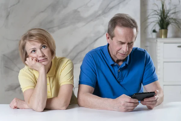 Elderly couple, senior woman wife looking unhappy bored while her retired man husband ignoring her, paying no attention, busy using his mobile cell phone. Smartphone, gadget addiction. Boring date
