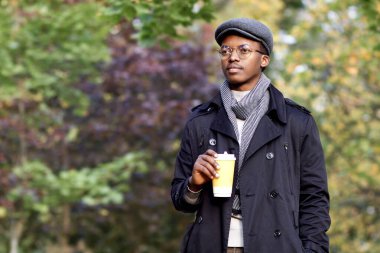Yakışıklı, ciddi siyahi bir adamın portresi, şapkalı, atkılı ve gözlüklü genç Afrikalı Afro Amerikalı bir adam Golden Autumn Park 'ta yürüyor. Güzel bir sonbaharda, bir fincan çay ya da kahveyle.