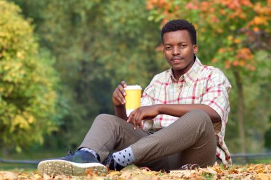Mutlu siyah adam, Afrikalı Afro-Amerikalı yakışıklı genç adam çimlerin üzerinde oturuyor, sonbaharda altın sonbahar parkında kahve içiyor yapraklara bakıyor, kameraya bakıyor.