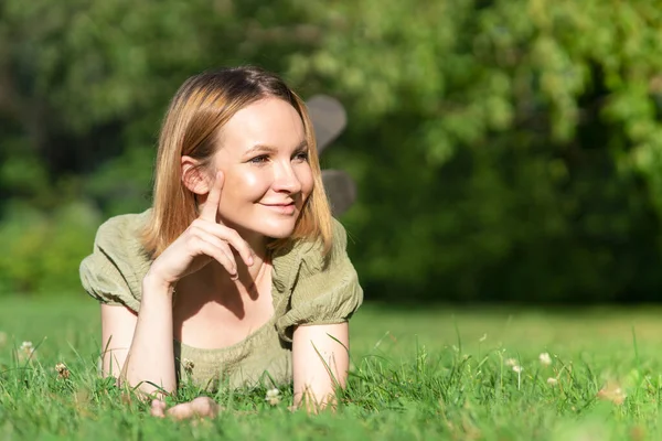 Beautiful happy positive girl, young cheerful woman is lying on her chest on green grass in the park or forest and smiling, enjoying walking, good sunny warm hot weather, looking away, copy space Royalty Free Stock Photos