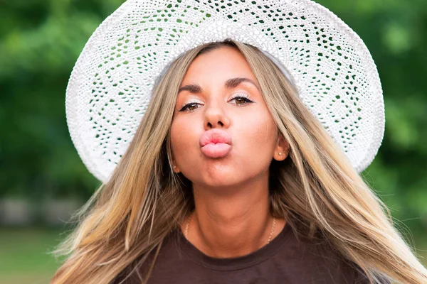 Close up beauty portrait of beautiful young woman, pretty attractive girl in wicker summer hat is kissing a camera, sending, blowing a kiss with big plump lips in a green park. Silicone female lips Stock Photo