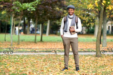 Mutlu siyahi üniversite öğrencisinin tam boy portresi ya da üniversite öğrencisi, gözlüklü, şapkalı, sırt çantalı ve kitaplı genç Afrikalı Afro Amerikalı sonbaharda Golden Autumn Park 'ta yürüyor, gülümsüyor.