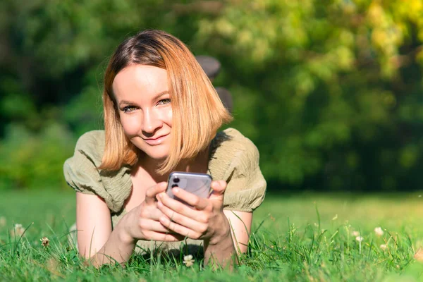 Retrato de menina bonita atraente, jovem feliz alegre mulher positiva está deitado no parque de verão na grama, sorrindo e olhando para a câmera com seu telefone celular, smartphone — Fotografia de Stock