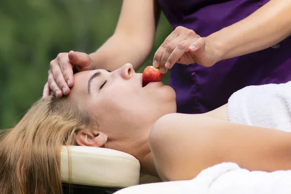 Professional masseur is feeding a beautiful woman with strawberry after procedure, massage. Beautiful relaxed young girl is lying on massaging table, eating berry outdoors. Health body care.