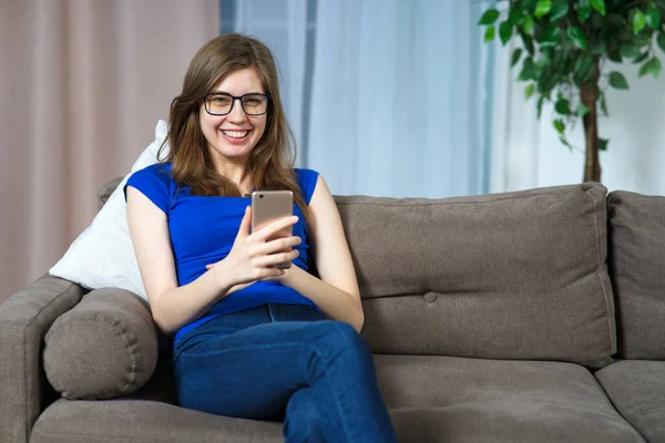 Retrato de menina adolescente feliz, jovem mulher alegre em óculos sentado no sofá na sala de estar em casa, rindo com seu telefone celular, smartphone na mão, olhando para a câmera, se divertindo — Fotografia de Stock