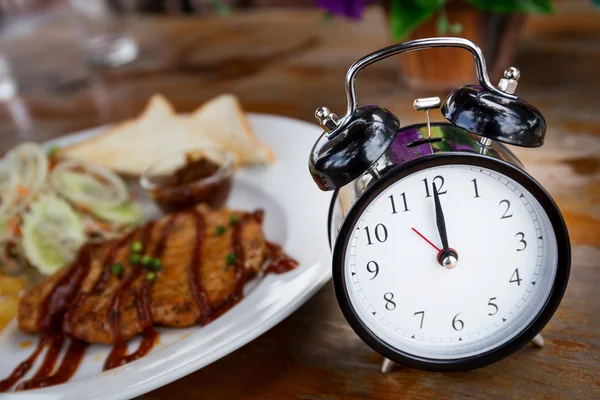 Uhr auf Holztisch mit Steak im Hintergrund, Mittagspause Stockbild