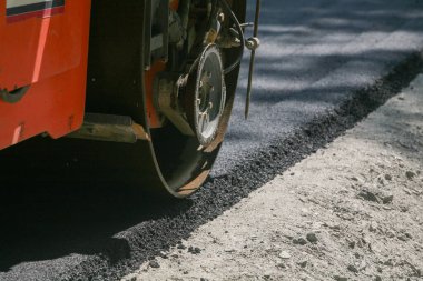 Turuncu bir yol silindirinin ağır çelik tekerleğinin inşaat sırasında yol yatağına kalın bir siyah asfalt şeridi bastırışının yakın görüntüsü.