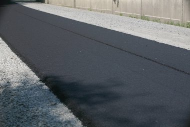 Uzun, yeni asfaltlanmış, koyu siyah asfalt bir yol yüzeyi daha hafif çakıllı bir omuz yanında, arka planda beton bir çit görünür.