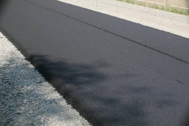 Uzun, yeni asfaltlanmış, koyu siyah asfalt bir yol yüzeyi daha hafif çakıllı bir omuz yanında, arka planda beton bir çit görünür.