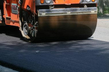 Bir inşaat sahasında yeni yerleştirilmiş siyah asfalt kaldırımı sıkıştıran turuncu bir tek davullu titreşimli silindir.