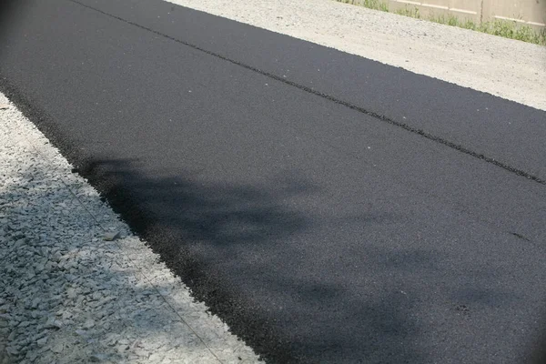 Uzun, yeni asfaltlanmış, koyu siyah asfalt bir yol yüzeyi daha hafif çakıllı bir omuz yanında, arka planda beton bir çit görünür.